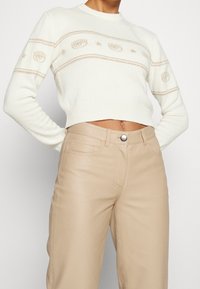 Pull court blanc avec motifs en forme d'étoiles et en forme de cils beiges, associé à un pantalon beige taille haute, dévoilant le ventre et les mains derrière le dos.