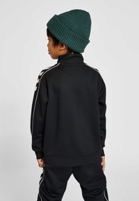 Chaqueta negra con cremallera y cuello alto, con rayas blancas en las mangas. Combinada con pantalones negros, lleva puesta una gorra de lana verde.