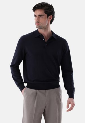 Mann trägt ein schwarzes langärmeliges Polohemd und beige plissierte Hosen, steht mit einer Hand in der Tasche und schaut leicht nach links.