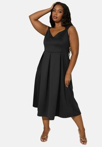 Robe noire sans manches en A avec un décolleté en V, jupe plissée et texture lisse. Associée à des sandales dorées à lanières.