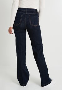 Jeans en denim bleu foncé évasés avec une taille haute, des coutures visibles et deux poches arrière. Les jeans ont une texture en denim lisse et un léger éclat.