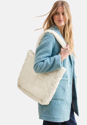 Borsa tote in avorio trapuntata con manici imbottiti, appoggiata su un cappotto in fleece blu testurizzato, con zip e motivo a quadrati sulla borsa.