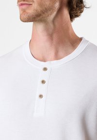 Valkoinen henley-paita, jossa on resorikuvio, pyöreä pääntie ja kolme ruskeaa nappia laitteen varrella. Yksinkertainen ja klassinen muotoilu.