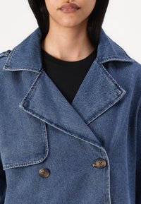 Denim jakna z dvojnim zapenjanjem, z obrobnim ovratnikom, kontrastnim šivanjem in dvema temnima gumboma, v srednje modrem odtenku z mehko teksturo.