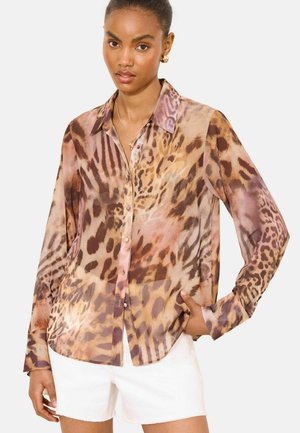 Žena v dlhých rukávoch, zapínanej na gombíky, s potlačou zvierat a golierom, a v bielych kraťasoch, pózuje s jednou rukou v vrecku na jednoduchom pozadí.