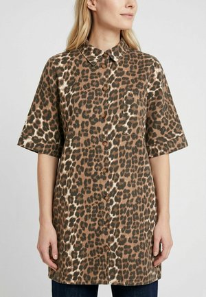 Femme portant une chemise boutonnée marron et noire à imprimé léopard avec manches courtes et une poche poitrine, debout devant un fond clair.