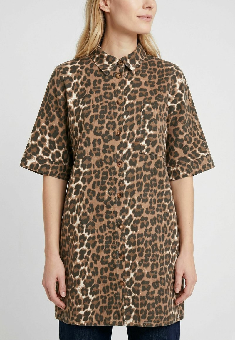 Femme portant une chemise boutonnée marron et noire à imprimé léopard avec manches courtes et une poche poitrine, debout devant un fond clair.