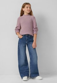 Jong meisje met lang haar, gekleed in een lila gebreid sweater, wijdvallende blauwe jeans, witte sneakers en staande met één hand in haar zak.