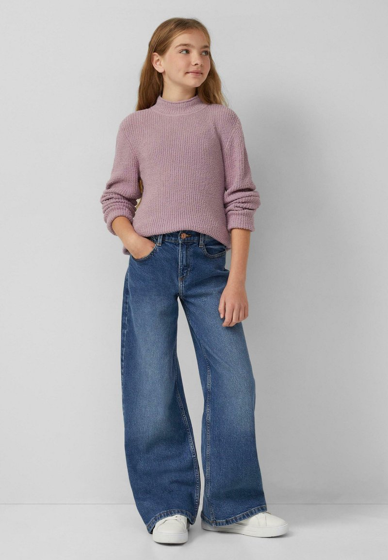 Jong meisje met lang haar, gekleed in een lila gebreid sweater, wijdvallende blauwe jeans, witte sneakers en staande met één hand in haar zak.