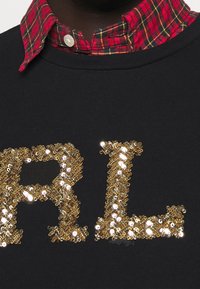 Sudadera negra con grandes letras de lentejuelas doradas "R" y "L." Cuello de camisa a cuadros rojo y negro visible debajo. Tela texturizada.