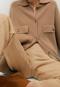 Cardigan a maglia beige con colletto, dotato di due tasche frontali e bottoni in legno, abbinato a pantaloni beige coordinati e stivali in suede marrone.