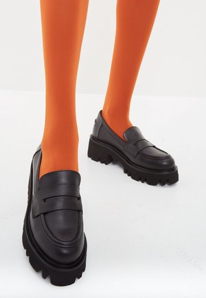 Ben iført lyse orange strømpebukser kombineret med sorte chunky platform loafers på en hvid overflade.