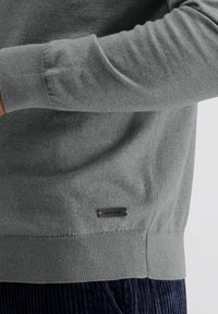 Grijze gebreide trui met een geribde halslijn en manchetten. Voorzien van een zwart label met "pierre cardin" aan de zijkant. Zachte textuur en casual pasvorm.