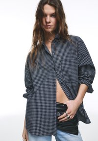 Kockovaná tmavosivá a čierna nadrozmerná košeľa na gombíky s vyhrnutými rukávmi, v kombinácii s čiernou koženou bundou a svetlomodrými džínsami.