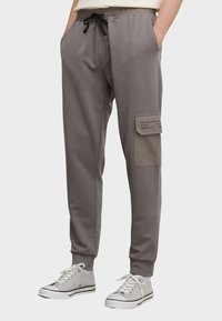 Graue Baumwoll-Jogginghosen mit elastischem Bund und schwarzem Kordelzug. Mit Seitentaschen und einer aufgesetzten Tasche am rechten Bein. Trägt sich gut mit Sneakers.