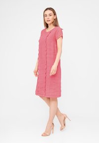 Robe en lin rose avec un col rond, des manches courtes et un détail festonné sur le devant. Associée à des sandales nude à talons hauts.