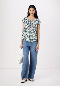 Haut multicolore à manches courtes avec des motifs floraux abstraits ; porté avec un jean bleu à jambe large et un sac à main blanc. Les chaussures sont de teinte claire.