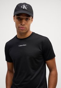 Czarna, krótkosleeve koszulka sportowa z teksturowanym wykończeniem i małym białym logo "Calvin Klein" na klatce piersiowej; zestawiona z czapką w kolorze czarnym.