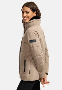 Beige puffer jacket with a high collar, black faux fur lining, and logo patch, featuring zippered side pockets and a quilted design.