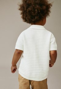 Vit kortärmad pikétröja med ett texturerat mönster, ribbad krage och mudd, kombinerad med ljusbruna byxor. Visad bakifrån.