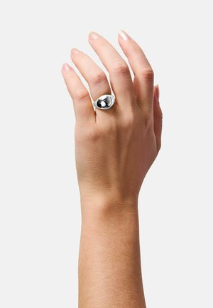 Skulpturel sølvring med en glat, rund form og en blank, reflekterende sort accent. Båret på en menneskehånd mod en ensfarvet baggrund.