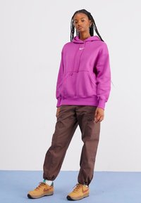 Μωβ υπερμεγέθη κουκούλα με μπροστινή τσέπη καγκουρό και κορδόνια, συνδυασμένη με καφέ cargo παντελόνια και κίτρινα αθλητικά παπούτσια. Απλό φόντο.