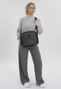 Zwarte ribbels laptop tas met een gladde voorpaneel, bovenhandvat en verstelbare schouderband, gedragen op de schouder tegen een lichte achtergrond.