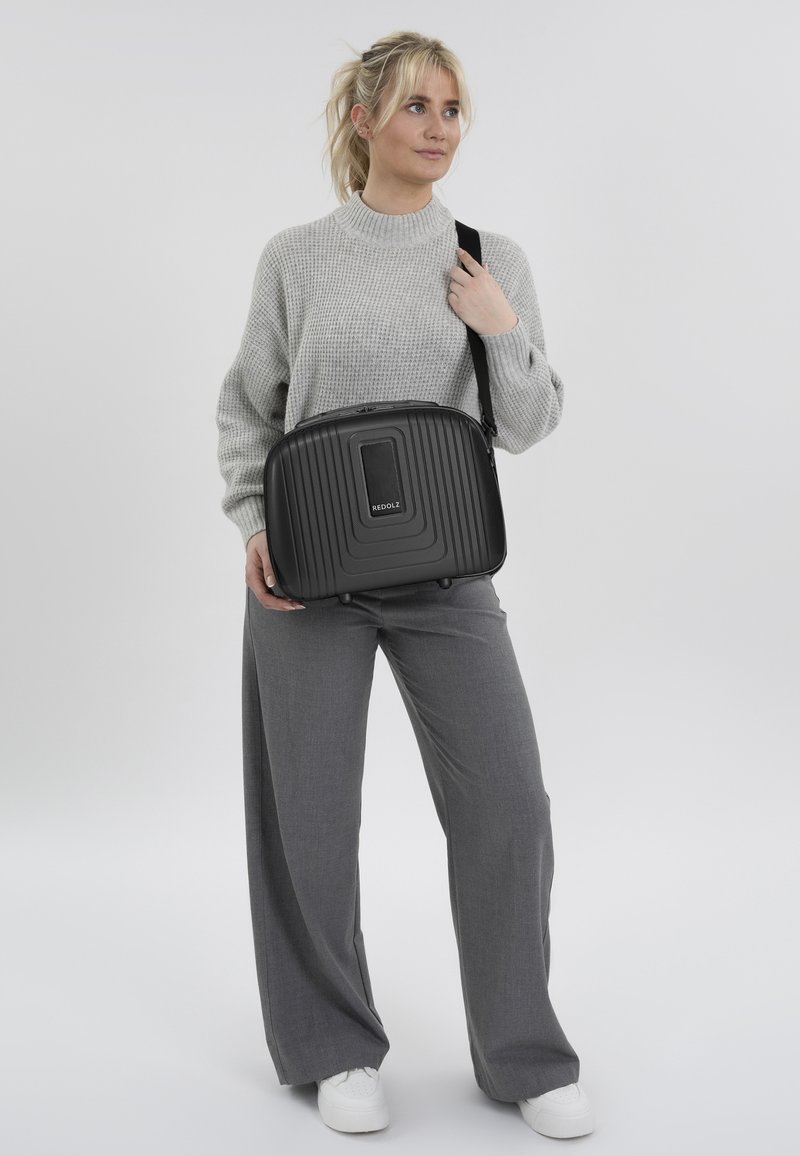 Zwarte ribbels laptop tas met een gladde voorpaneel, bovenhandvat en verstelbare schouderband, gedragen op de schouder tegen een lichte achtergrond.