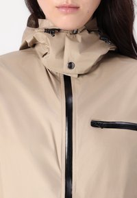 Beżowa wodoodporna kurtka z wysokim kołnierzem i regulowaną kapturem. Posiada czarny zamek błyskawiczny z przodu oraz zamykaną na zamek kieszeń na piersi. Gładka tekstura.