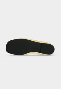Semelle noire en caoutchouc d'une chaussure plate beige avec une adhérence texturée aux zones du talon et de l'avant-pied, présentée sur un fond clair uni.
