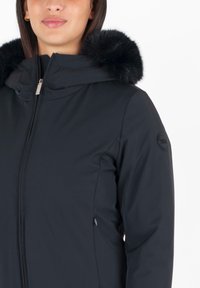 Zwarte waterdichte jas met een afneembare, fluffy zwarte bontkap. Voorzien van een voorrits en zijzakken voor opbergruimte.