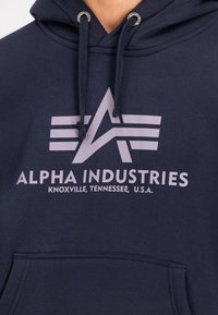 Felpa blu navy realizzata in tessuto morbido con tasca frontale. Sul petto è presente un logo grigio chiaro e la scritta "ALPHA INDUSTRIES" con strisce.