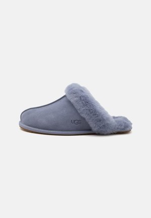 Απαλή γαλάζιο-γκρι παντόφλα τύπου slip-on με επένδυση και διακόσμηση από πλούσια γούνα, χαμηλό τακούνι και λογότυπο UGG στο πλάι.