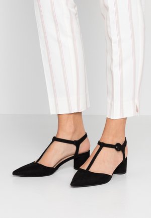 Femme portant des escarpins en daim noir à bride en T avec talons carrés, associés à un pantalon blanc à fines rayures verticales rouges.