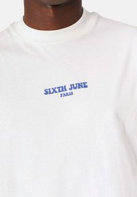 Hvid bomulds-t-shirt med blå tekst "SIXTH JUNE PARIS" i fed skrift, med rund halsudskæring og standard pasform.