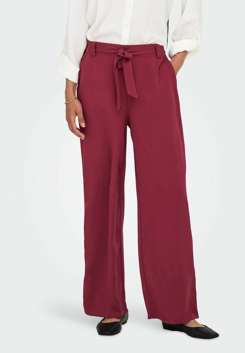 Pantalon large bordeaux en tissu léger, avec taille nouée, poches latérales et texture lisse. Associé à des ballerines noires.