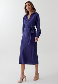 Abito avvolgente a maniche lunghe di un profondo blu navy con una cintura legata in vita, caratterizzato da un colletto e un tessuto liscio. Indossato con sandali con tacco color pelle.
