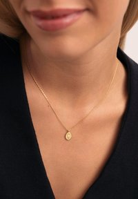 Collar de oro con una cadena delgada y un colgante ovalado que presenta un diseño central. El fondo es oscuro, lo que resalta la joyería.