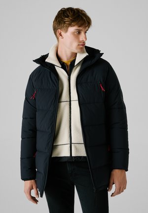 Zwarte pufferjas met capuchon, voorzien van rode ritsdetails, gedragen over een witte fleece binnenlaag. Gelaagd stof en gestikte ontwerp.