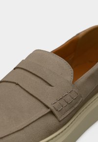 Μπεζ μοκασίνι από σουέντ με απαλή υφή, με ραμμένη λεπτομέρεια στην κορυφή και σόλα σε κρεμ απόχρωση. Εσωτερικά είναι πορτοκαλί.