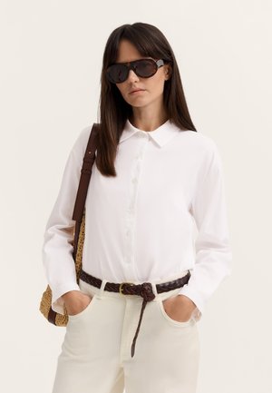 Frau mit dunkler Sonnenbrille, weißem Hemd mit Knöpfen, weißer Hose mit geflochtenem braunem Gürtel und tragender gewebter Schultertasche.