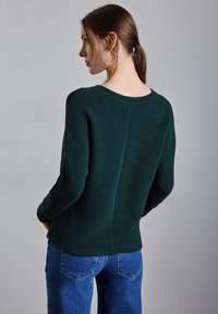 Maglione verde scuro con texture, scollatura rotonda e maniche lunghe, abbinato a jeans blu. Il retro presenta un dettaglio di cucitura verticale.