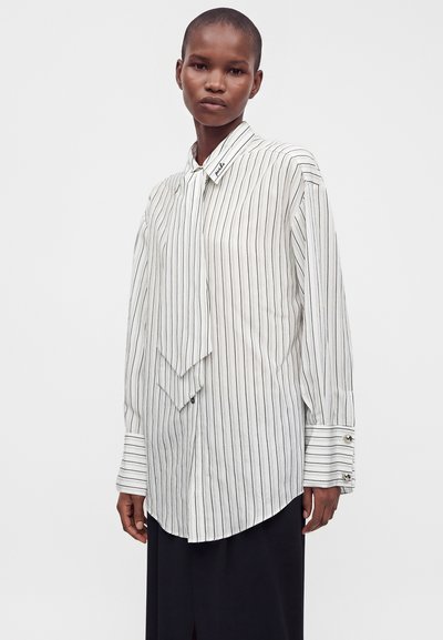 Personne portant une chemise blanche à manches longues avec de fines rayures verticales noires et de larges poignets, associée à une jupe foncée sur un fond uni.