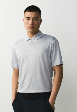 Junger Mann mit kurzen Haaren, der ein hellgraues Poloshirt mit Kragen und dunkle Hosen trägt, die Hände in den Taschen, vor einem einfarbigen Hintergrund.