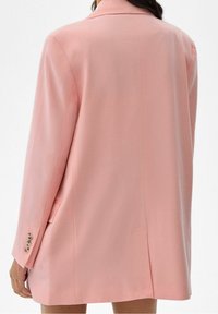 Blazer rosa su misura con una texture liscia, rever a rotondo e bottoni decorativi sulle maniche. Presenta una fessura centrale sul retro per maggiore mobilità.