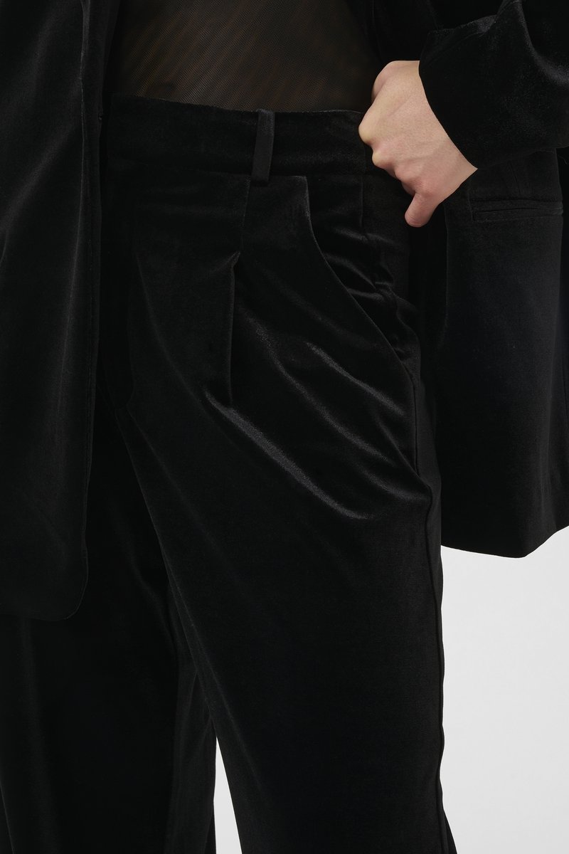 Schwarze Samthose mit glatter Textur, ausgestattet mit Falten und Seitentaschen. Kombiniert mit einem transparenten schwarzen Oberteil unter einem passenden Blazer.