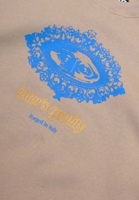Felpa beige con grafica blu raffigurante un design ornamentale con un motivo a occhio e testo giallo che recita "inter's quality" e "Forged in Italy."