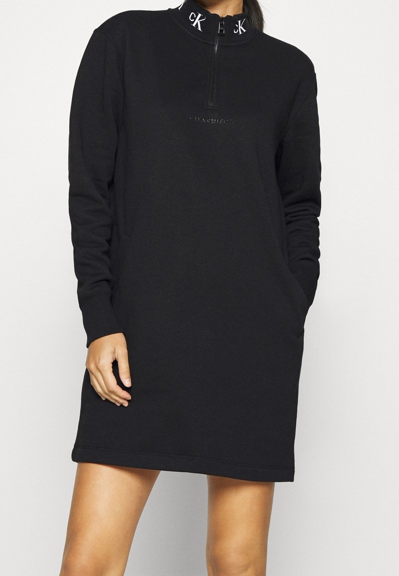 Femme portant une robe-pull noire à manches longues avec un col zippé affichant les lettres blanches "cK". Mains détendues, une dans la poche.