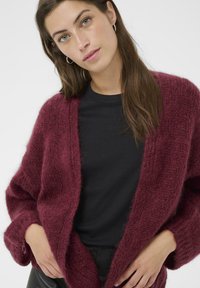 Burgundowa sweter z dzianiny z otwartym przodem, z luźnymi rękawami i miękkim, teksturowanym wykończeniem. Połączony z czarną bluzką.