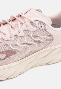 Zapatilla deportiva de color rosa claro con parte superior de malla, patrones texturizados, detalles ondulados en blanco y una suela gruesa de color crema. Cuenta con cordones y orificios para ventilación.